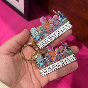 May include: Two Birmingham, Alabama, skyline keychains with gold-colored key rings. The keychains feature a colorful cityscape design with the city name in white. The background is a bright pink.