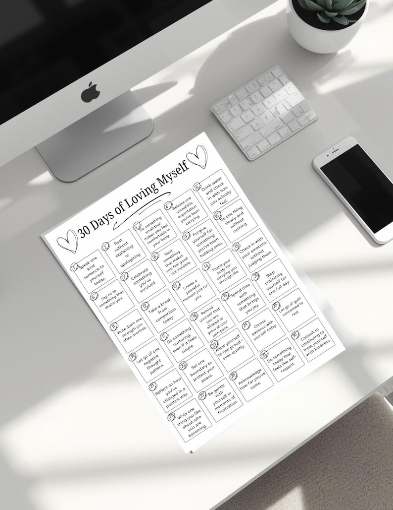 May include: A white paper titled "30 Days of Loving Myself" with a list of self-care prompts. The paper is on a white desk with a computer, keyboard, and phone. The prompts include "Speak one kind sentence to yourself today" and "Commit to treating yourself with kindness."