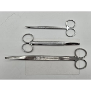 Könnte beinhalten: Drei chirurgische Scheren aus Edelstahl in verschiedenen Größen werden gezeigt. Die Scheren haben Fingerlöcher und scharfe Klingen. Das größte Paar hat eine gerade Klinge, während die anderen beiden gebogene Klingen haben. Die Scheren liegen auf einer weißen Oberfläche.
