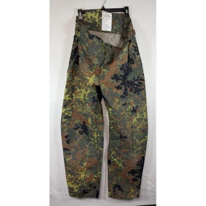 May include: Camouflage pattern pants in shades of green, brown, and black, hanging from a black hanger. The pants feature suspender straps and a tag. The fabric has a mottled design, typical of military-style clothing.