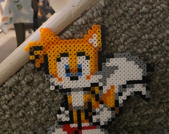 May include: A pixel art bead sprite of the video game character Tails from the Sonic the Hedgehog franchise. The character is yellow, white, and blue with a red stripe on his shoes. The bead sprite is on a gray carpet.