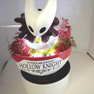 May include: A decorative display featuring a white Hollow Knight character figure with dark brown accents, positioned inside a bowl-shaped base. The base is red and white with the text HOLLOW KNIGHT printed on the front, filled with moss and LED lights.