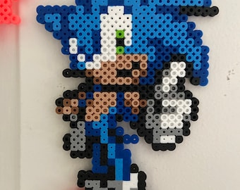 May include: A blue and white pixel art character of Sonic the Hedgehog, made with colorful beads. The character is standing with his arms at his sides and his legs spread apart. He is wearing red shoes with white laces.