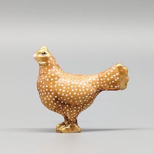 Könnte beinhalten: Eine kleine Keramikfigur eines braun-weiß gefleckten Huhns. Das Huhn steht auf seinen beiden Beinen mit dem Kopf nach oben geneigt. Die Figur ist mit goldenen Akzenten bemalt.