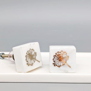 May include: Two white square cabinet knobs with a gold dandelion design. The knobs are on a white surface.