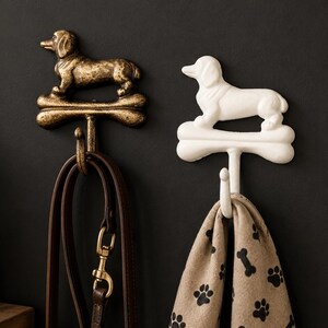 May include: Two wall-mounted hooks shaped like dachshunds standing on bones. One hook is finished in a distressed metallic bronze, holding a brown leather leash. The other hook is matte white, holding a beige fabric with black paw and bone prints.