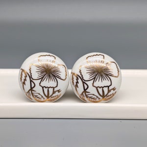 Op de afbeelding: Twee witte keramische ladeknopjes met gouden bloemmotieven. De knoppen zijn rond en hebben een reliëfdesign.