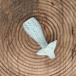 May include: White ceramic mermaid tail figurine with a textured, scaly design. The tail is curled up and the fin is pointed. The figurine is on a wooden background.