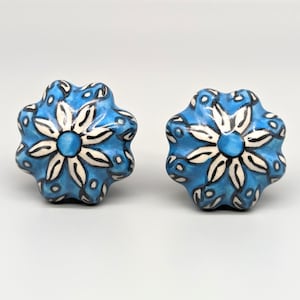 Può includere: Due pomelli per armadi in ceramica blu con un motivo floreale bianco. I pomelli hanno una forma a stella con un centro rialzato.