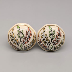 Op de afbeelding: Twee keramische kastknoppen met een zeshoekige vorm. De knoppen zijn wit met een gouden rand en een bloemmotief in tinten groen, oranje en geel.