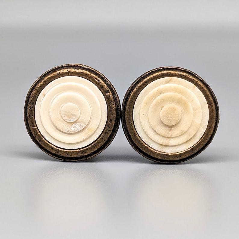 May include: Two round, decorative knobs with a concentric circle design. The knobs are made of white bone with a bronze metal rim.