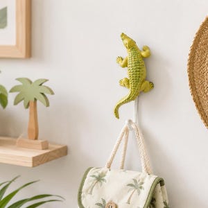 Puede incluir: Un gancho de pared con forma de caimán texturizado de color verde lima. El caimán está colocado verticalmente con la cabeza hacia arriba y la cola hacia abajo, sirviendo como un gancho funcional para colgar un bolso pequeño con palmeras.