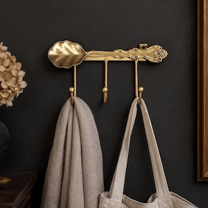 May include: A gold-toned wall-mounted hook rack shaped like a vintage spoon with ornate handle detailing. The horizontal bar features three downward-curving hooks, with a beige fabric scarf and a light-colored canvas tote bag hanging from them.