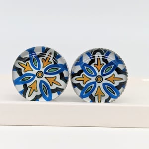 May include: A pair of round, blue and white glass earrings with a silver trim. The earrings feature a geometric pattern with yellow accents.