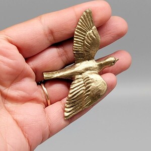 May include: A detailed, gold-colored bird figurine in mid-flight. The wings are outstretched, showcasing intricate feather detailing. The bird is a decorative item, possibly a brooch or pendant, with a vintage or antique aesthetic.