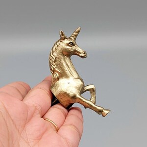 May include: A gold-colored unicorn figurine with a spiraled horn, mane, and raised front leg. The mythical creature is detailed with a textured surface and is held in a hand against a gray background.