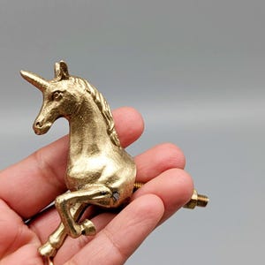 May include: A gold-colored unicorn figurine with a detailed mane and horn. The unicorn is posed in a rearing position, with its front legs raised. The figurine has a screw attached to the back, suggesting it is a decorative cabinet knob or pull.