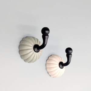 Puede incluir: Dos ganchos decorativos de pared. Cada gancho presenta un gancho de metal negro curvado unido a una base de cerámica festoneada. Una base es de color gris claro, la otra es rosa claro. Los ganchos están montados en una pared azul claro.