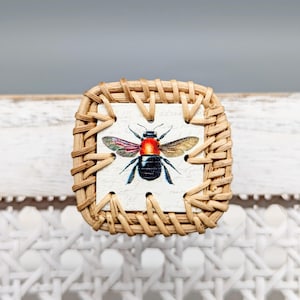 May include: A square wicker coaster with a white tile featuring a vintage illustration of a bee with red and black markings.