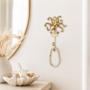 May include: A gold-toned metal wall hook shaped like an octopus with multiple curved tentacles extending outwards. The hook features a vertical stem ending in a rounded knob, currently holding a single strand of white beaded jewellery with gold accents.