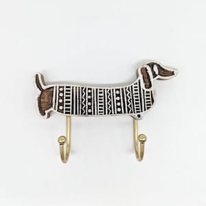 May include: White wooden dachshund shaped wall hook with a black and white geometric pattern. The hook has two gold metal hooks.