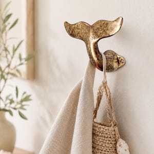 Può includere: Un gancio a forma di coda di balena color bronzo montato su una parete bianca. Un asciugamano di lino beige e una piccola borsa intrecciata con una conchiglia sono appesi al gancio. Il gancio ha una finitura strutturata.