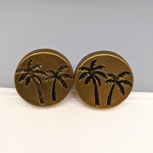 May include: Two round, bronze-colored plugs with a palm tree design. Each plug features two palm trees in a dark, contrasting color, set against a circular background. The plugs are likely for ear piercings.