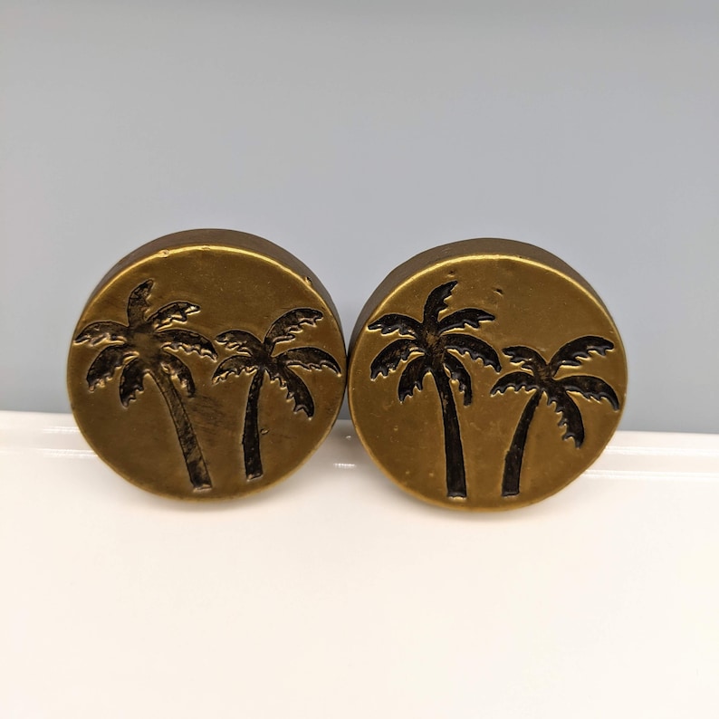 May include: Two round, bronze-colored plugs with a palm tree design. Each plug features two palm trees in a dark, contrasting color, set against a circular background. The plugs are likely for ear piercings.