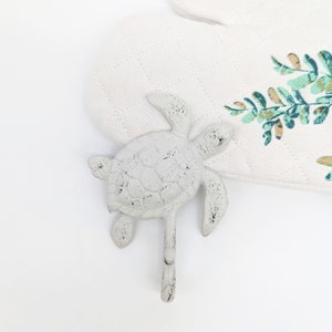 Peut inclure: Un crochet en forme de tortue en fonte gris clair vieilli. Le crochet a la forme d'une tortue de mer avec un seul crochet s'étendant vers le bas. Un gant de cuisine matelassé blanc avec un imprimé botanique est en arrière-plan.
