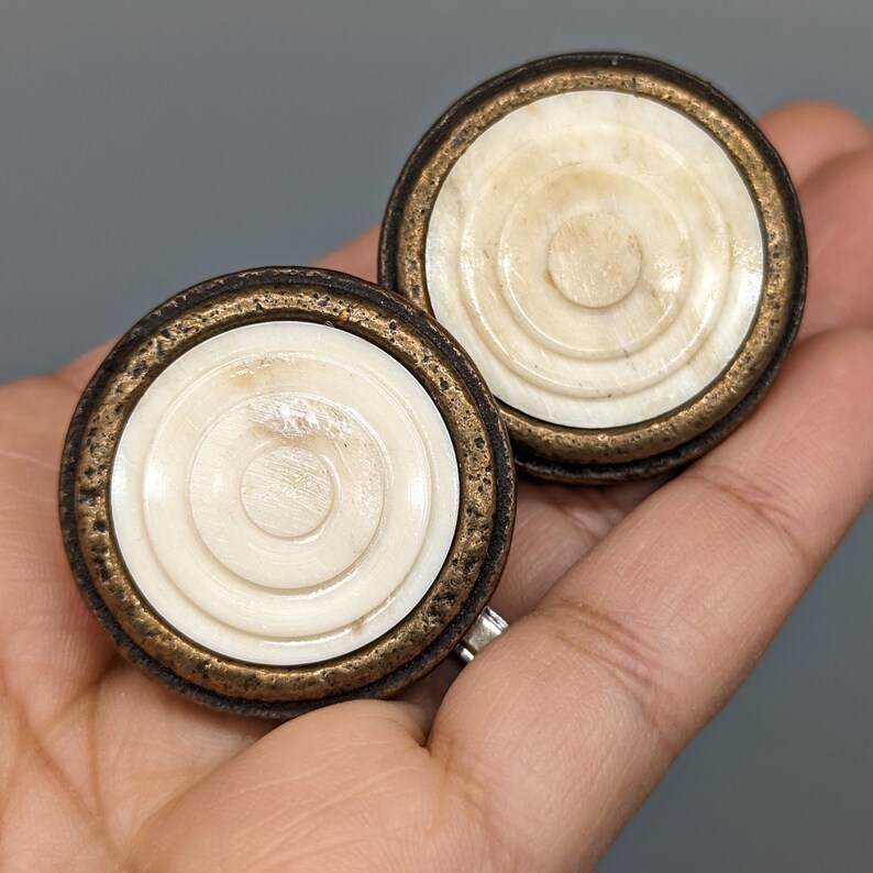 May include: Two round, decorative cabinet knobs with a brass finish and a white, concentric circle design.