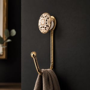 May include: A gold-toned metal wall hook featuring a decorative oval ceramic knob with a black, white, and tan geometric pattern. The hook has a curved lower arm with a ball-shaped tip, and a grey fabric item is draped over the lower section.