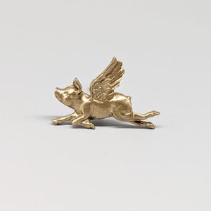 May include: A gold-colored figurine of a pig with wings, in mid-flight. The pig is detailed with visible ears, snout, and legs. The wings are intricately designed, adding to the whimsical nature of the piece. The figurine is set against a plain white background.