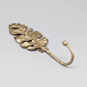 Puede incluir: Un gancho de pared de metal dorado con forma de hoja. El gancho tiene un extremo curvado y un diseño decorativo de hoja. El gancho está diseñado para colgar objetos y añade un toque de decoración inspirada en la naturaleza.