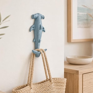 May include: A blue metal hammerhead shark wall hook. The hook is fixed to a white wall and supports a woven tote bag with rope handles. This decorative hook is suitable for coastal or nautical themed interiors.