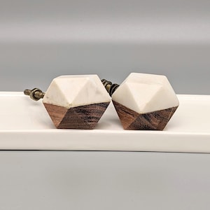 May include: A pair of geometric cabinet knobs. Each knob has a white upper section and a dark brown wooden base. The hardware is brass-coloured. The knobs are presented on a white surface against a grey background. The knobs are suitable for furniture.