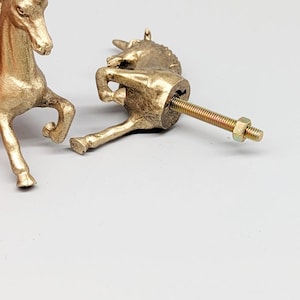 May include: Two gold-colored unicorn cabinet knobs. One knob is fully assembled with a screw and nut. The other unicorn knob is partially visible. The knobs are made of metal and have a detailed design. The background is a light gray color.