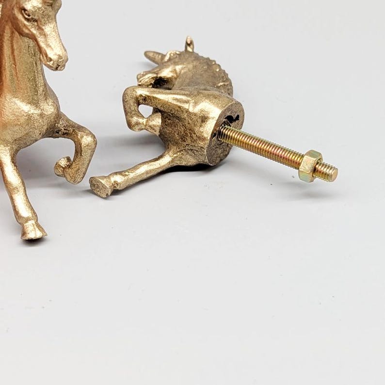 May include: Two gold-colored unicorn cabinet knobs. One knob is fully assembled with a screw and nut. The other unicorn knob is partially visible. The knobs are made of metal and have a detailed design. The background is a light gray color.