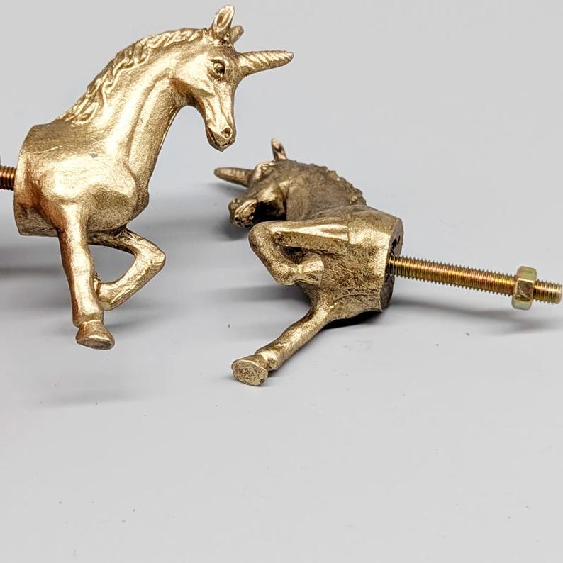 May include: Two gold-colored unicorn cabinet knobs. One knob is upright, the other is lying down. Both have a threaded bolt for installation. The knobs are made of a metallic material and have a detailed, textured finish.
