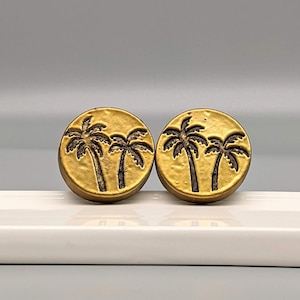 May include: Two round, gold-colored earrings with a palm tree design. Each earring features three palm trees in a dark brown color. The earrings are displayed on a white surface against a light gray background.
