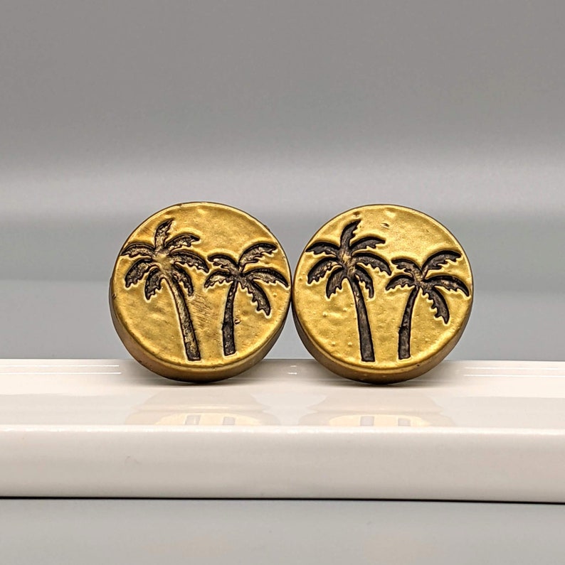 May include: Two round, gold-colored earrings with a palm tree design. Each earring features three palm trees in a dark brown color. The earrings are displayed on a white surface against a light gray background.