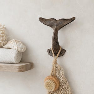 May include: A dark brown, cast iron wall hook shaped like a whale tail. The hook features a textured, weathered finish and two mounting holes at the base. A natural jute rope is looped around the hook, holding a small wooden bath brush.