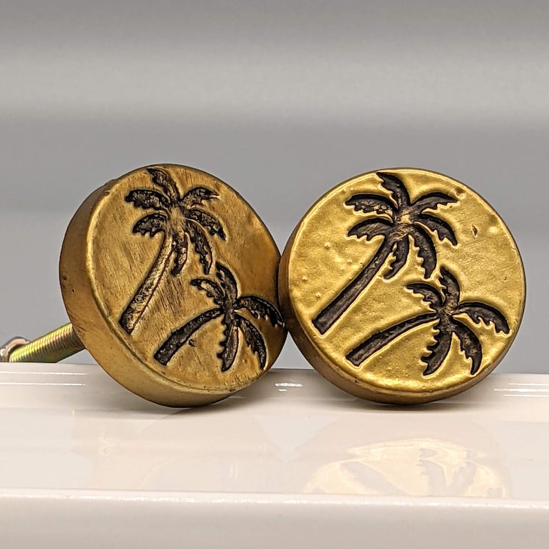 May include: Two gold-colored cabinet knobs with a palm tree design. Each knob features a circular face with a textured surface and a dark brown palm tree silhouette. The knobs are mounted on a white surface.