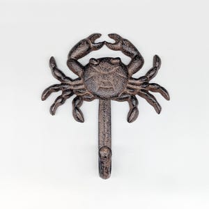 May include: A cast iron crab-shaped wall hook in a rustic brown finish. The hook features a detailed crab design with outstretched claws and multiple legs. The hook is mounted on a vertical stem with a curved hook at the bottom, perfect for coastal decor.