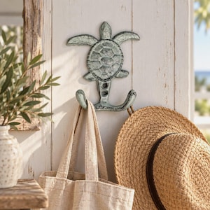 Peut inclure: Un crochet mural en forme de tortue de mer avec une finition vieillie vert clair et gris foncé. Le crochet présente une forme de tortue centrale avec deux bras incurvés s'étendant vers le bas, pour suspendre un sac fourre-tout en toile beige et un chapeau de paille.