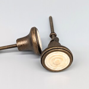 May include: Two antique brass cabinet knobs with white ceramic centers. The knobs have a circular design with concentric circles.