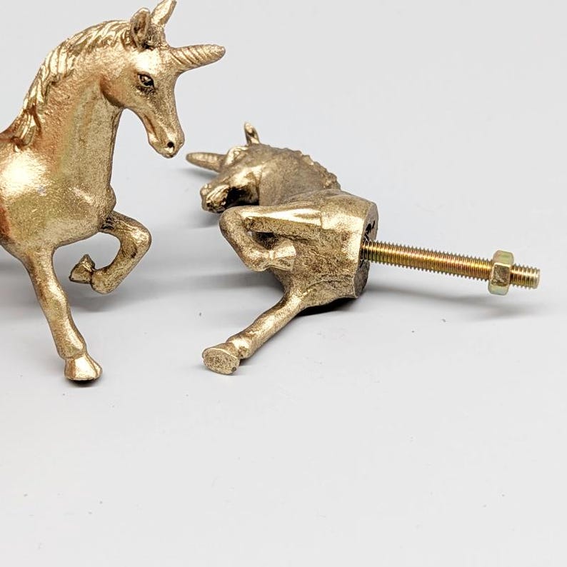 May include: Two gold-colored unicorn cabinet knobs. One unicorn is standing, the other is lying down with a threaded bolt attached. The knobs are made of metal and have a detailed, textured surface. The background is a plain, light gray.