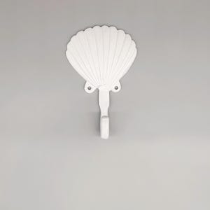 May include: A white seashell-shaped wall hook. The hook is made of a solid material and features a detailed shell design. The hook is designed to hang items and adds a coastal touch to any space.