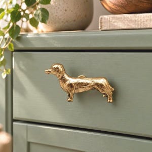 May include: A gold-toned metal drawer pull shaped like a standing dachshund dog. The hardware features a textured, metallic finish and is mounted horizontally onto the front of a sage green wooden drawer, serving as a functional handle.