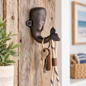 May include: A dark brown cast iron wall hook shaped like a whale, mounted on a vertical piece of weathered wood. The whale's tail curves upwards to form the hook, which holds a gold-toned metal keyring with keys and a beige rope lanyard.