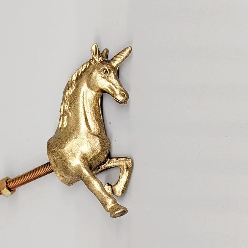 May include: A gold-colored unicorn figurine with a spiraled horn, raised front legs, and a copper-colored threaded rod attached to its body. The unicorn is set against a plain, light gray background.